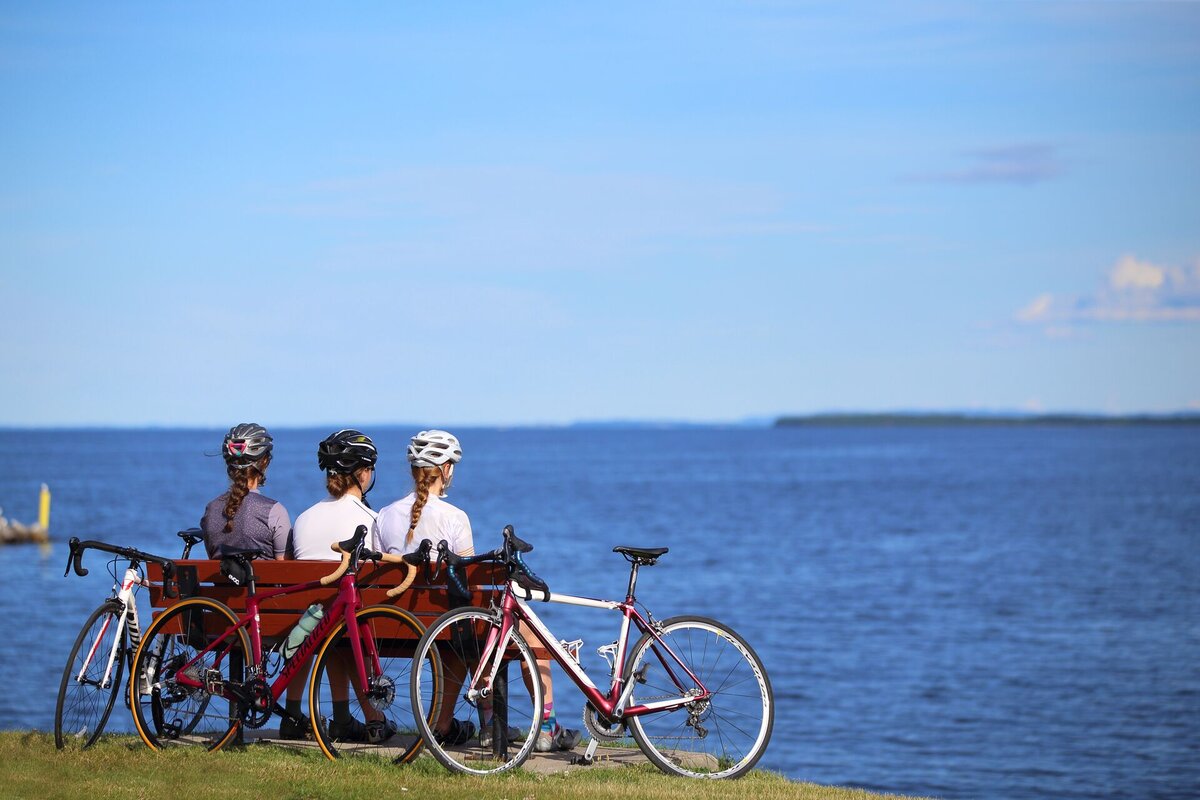 Three women sitting on a bench with their bikes, contemplating Lac-Saint-Jean